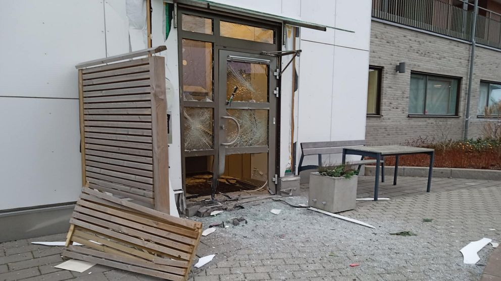 Skadad entré med spruckna fönster och en trasig dörr, där en del av fasaden har lossnat. Det ligger glas och skräp på marken, och en liggande träpanel är synlig vid sidan av dörren. I bakgrunden skymtar en grön växt i en kruka och byggnadens väggar.