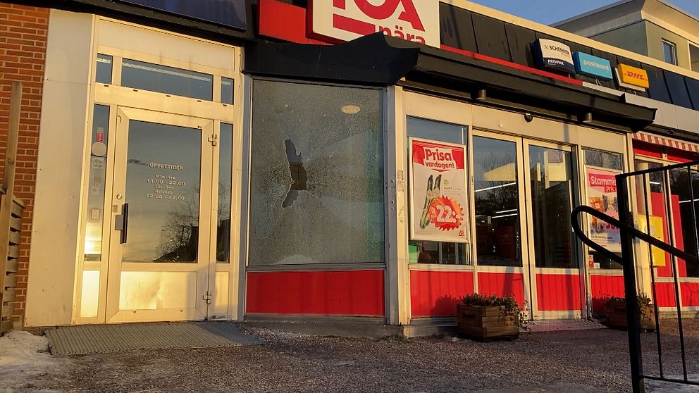 Krossad fönsterruta vid ingången till en Ica-butik med synliga skyltar och reklam. Butikens dörr är öppen och det finns snö på marken framför.