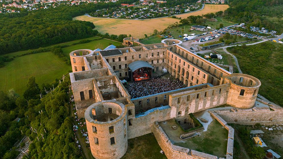 Fågelperspektiv av en historisk ruin med en konsert pågår inuti. Området är omgivet av grönska och fält, med en kustlinje i bakgrunden. Många människor samlas för att se uppträdandet i ruinen.