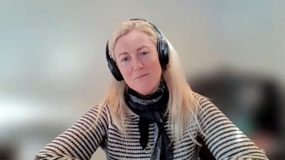 En blond kvinna med hörlurar, randig tröja och en knuten sjal runt halsen. Bakgrunden är suddig.