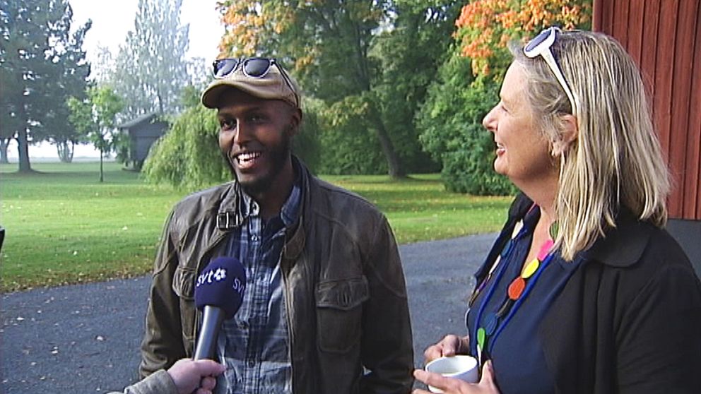 yngre man och medelålders kvinna intervjuas utomhus, båda glada och ler