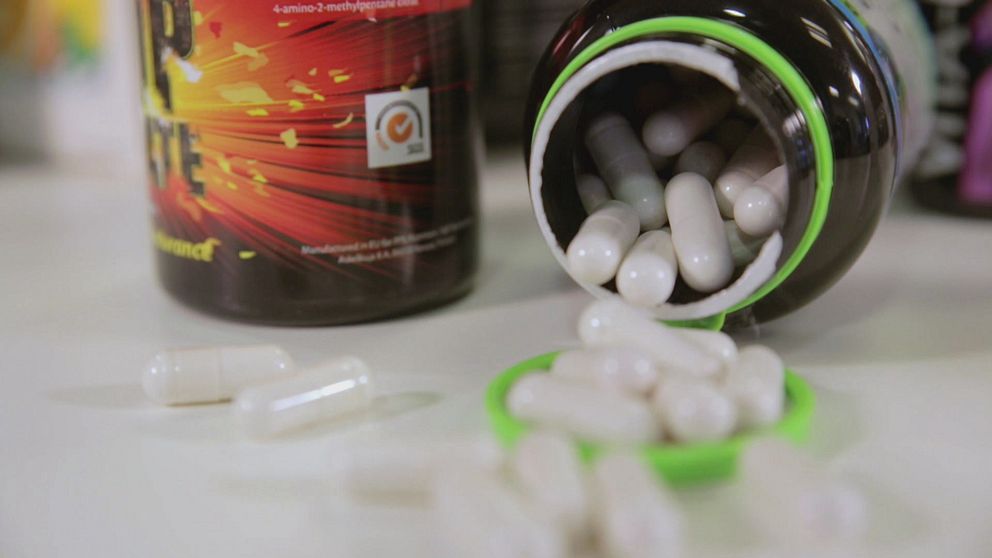 En rad pillerburkar syns suddigt i bakgrunden. I förgrunden till höger i bild ligger en öppen burk med vita piller som trillat ut över bordet.