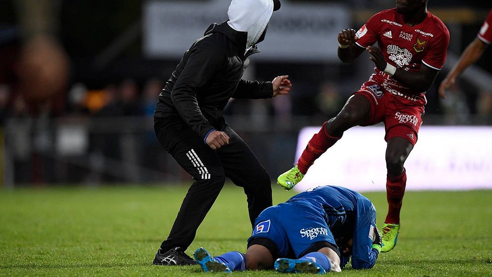 Maskerad man på en fotbollsplan. En spelare ligger på marken, en annan är på väg mot den maskerade mannen.