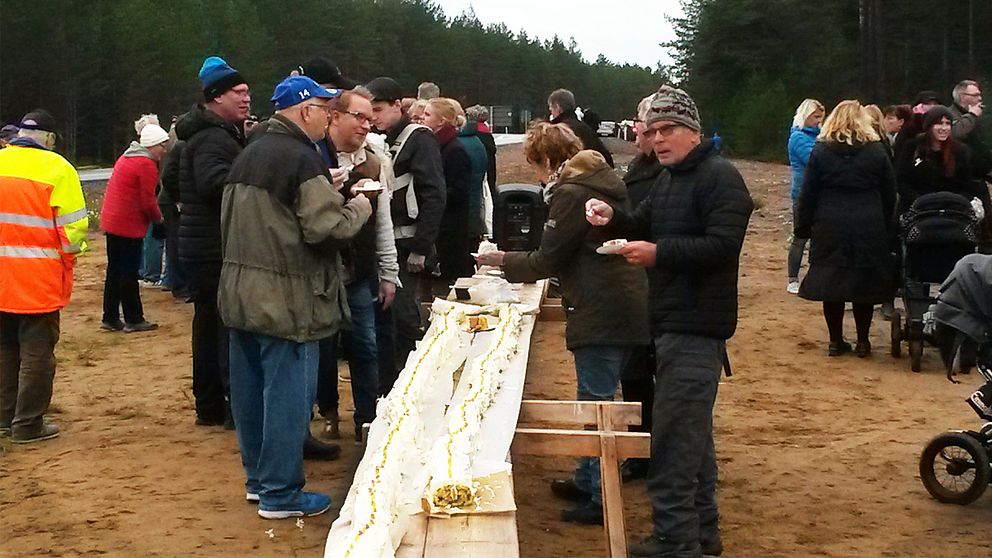 En lång rulltårta ligger på ett avlångt provisoriskt bord. Människor äter tårta. De är utomhus. Höga granar syns i bakgrunden.