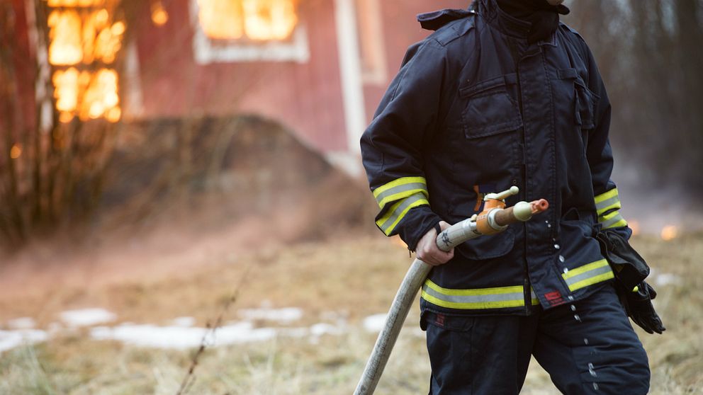 Brandman håller i brandslang framför brinnande hus