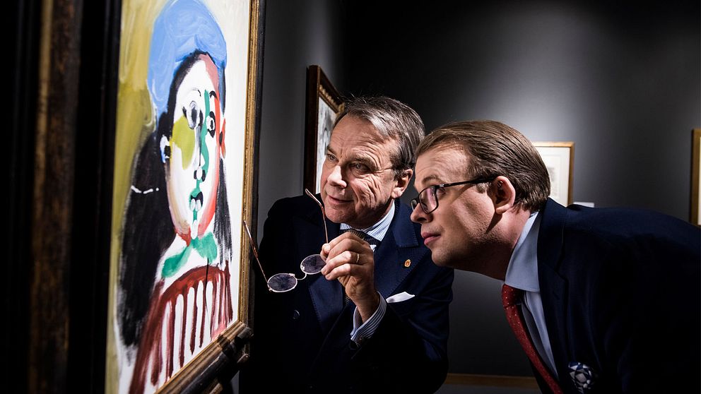 Till vänster i bild hänger en målning på en vägg. Två män betraktar bilden på nära avstånd. Mannen närmast tavlan håller sina glasögon i handen.