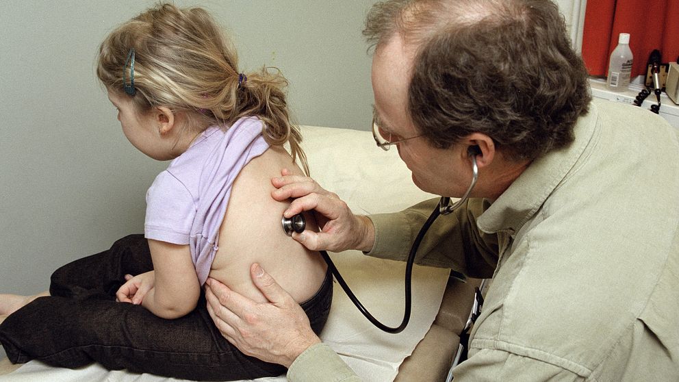 En barnläkare undersöker en flicka i 3-års ålder på ett undersökningbord. Flickan sitter upp.