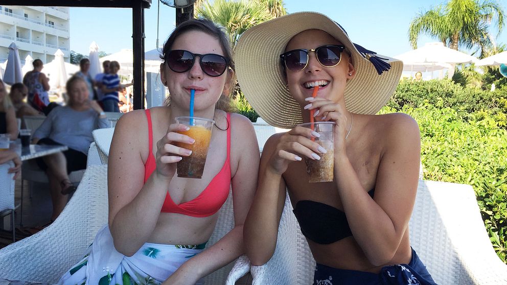 Två unga tjejer håller upp en varsin plastmugg med dryck och sugrör. De har solglasögon på sig. Det är soligt väder och palmer syns i bakgrunden. De är glada.