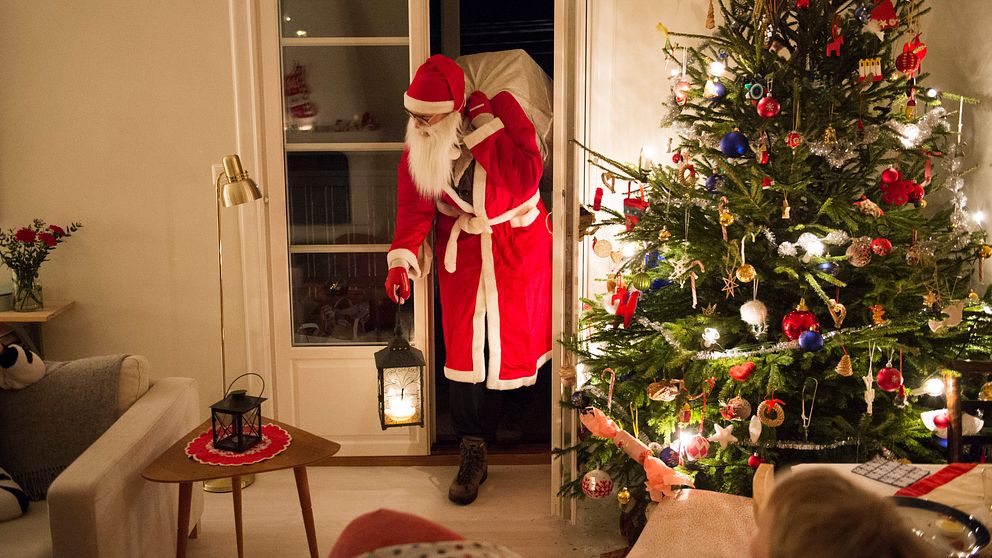 En jultomte är på väg in genom en altandörr, med en lykta i handen och en säck på ryggen. I rummet står en klädd julgran.