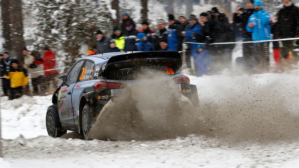 En rallybill sladdar i en kurva och kör upp brun snö. Vid sidan står publik och tittar på.