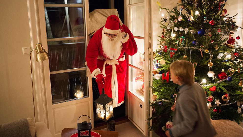 Jultomten kommer in genom altandörren. En pojke möter honom. I bakgrunden står julgranen.