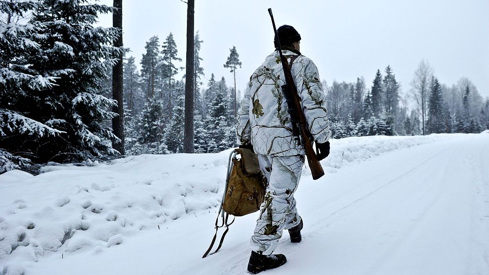 En bild på en jägare i kamouflagekläder med geväret på ryggen. Han går längs en väg i snölandskap.