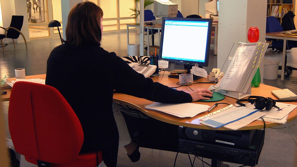 Kvinna sitter på en röd kontorsstol vid en arbetsplats och tittar mot en datorskärm, har högra handen på datormusen. På skrivbordet ligger papper och penna samt en telefon och hörlurar.