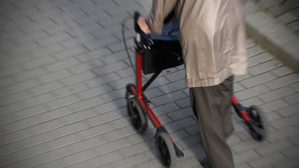 äldre person med brun kappa går med röd rollator