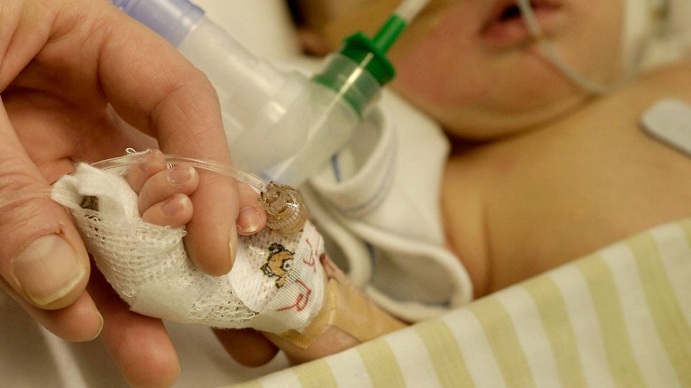 En vuxen hand håller i ett litet barns bandagerade hand på sjukhus. Barnet har slangar i näsan.