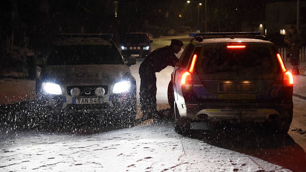 Två polisbilar står på en väg i mörker och snö, en polisman pratar med föraren i ena bilen.