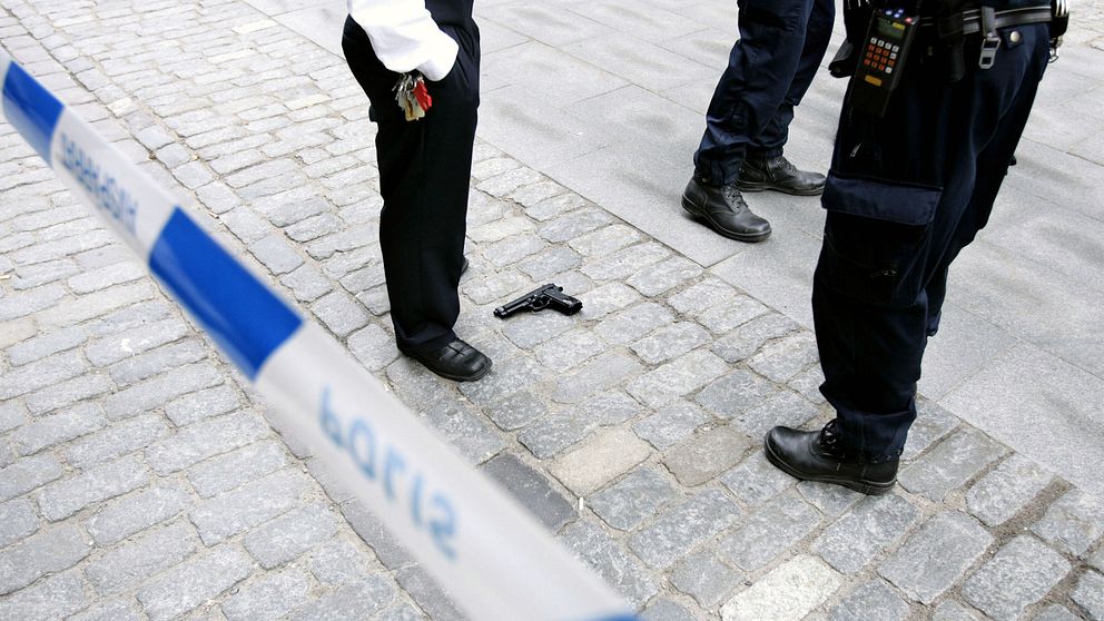 Blå-vitt polisavspärrningsband. Pistol på marken. Man ser vakter/polisers ben.