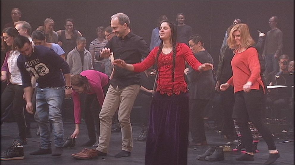 Kvinna i röd och violett romsk klädedräkt dansar med ett tjugotal amatörer på Folkoperans scen