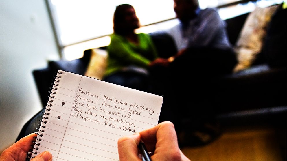 Bild över en rättspsykiater som skriver på ett skrivblock. I bakgrunden syns ett par som pratar om något.