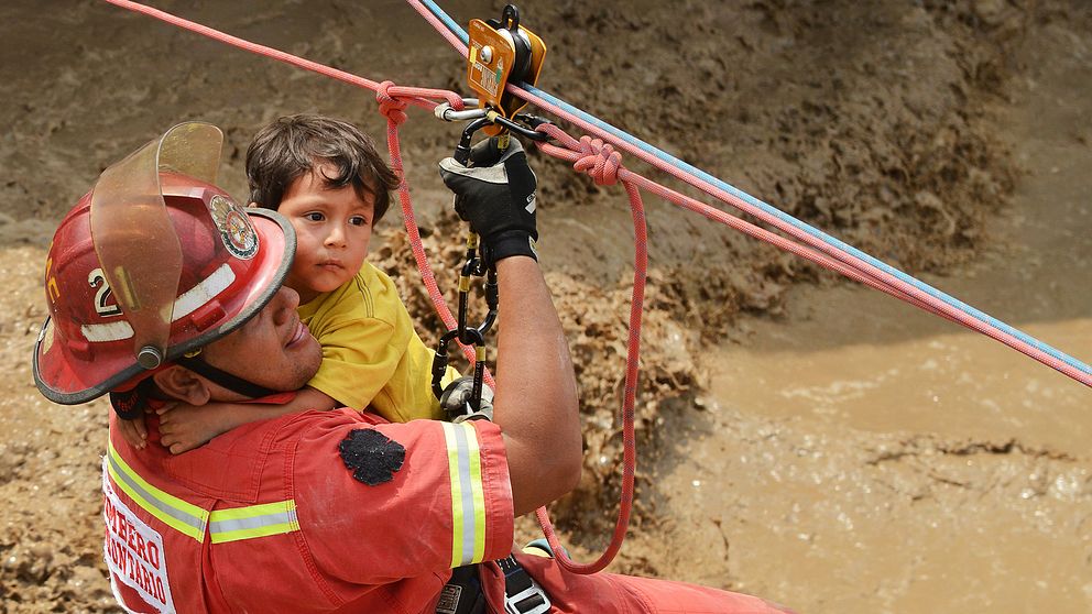 En liten pojke får hjälp av räddningstjänst att ta sig över en våldsamt forsande vattenmassa i Huachipa, öster om huvudstaden Lima.