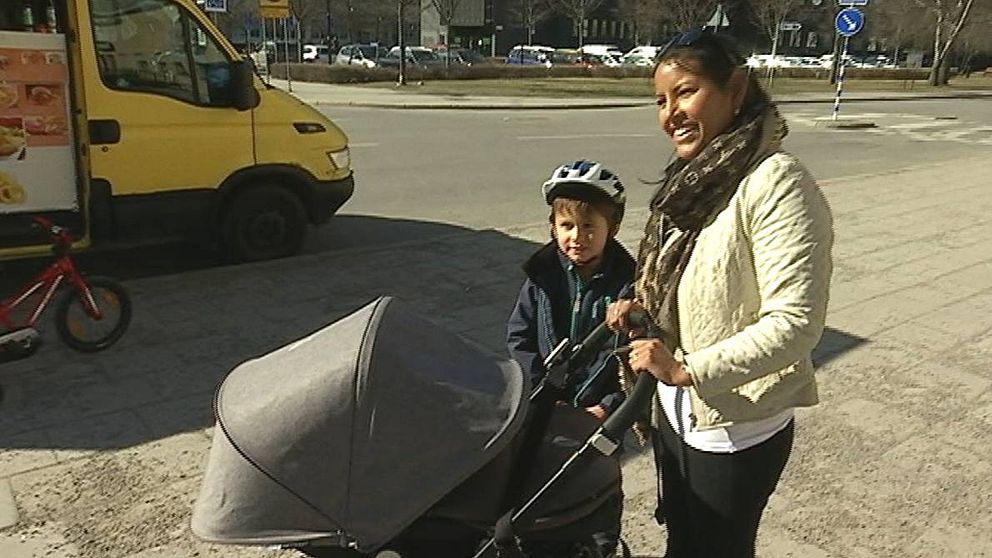 En kvinna står på trottoar med en barnvagn och ett litet barn med cykelhjälm.