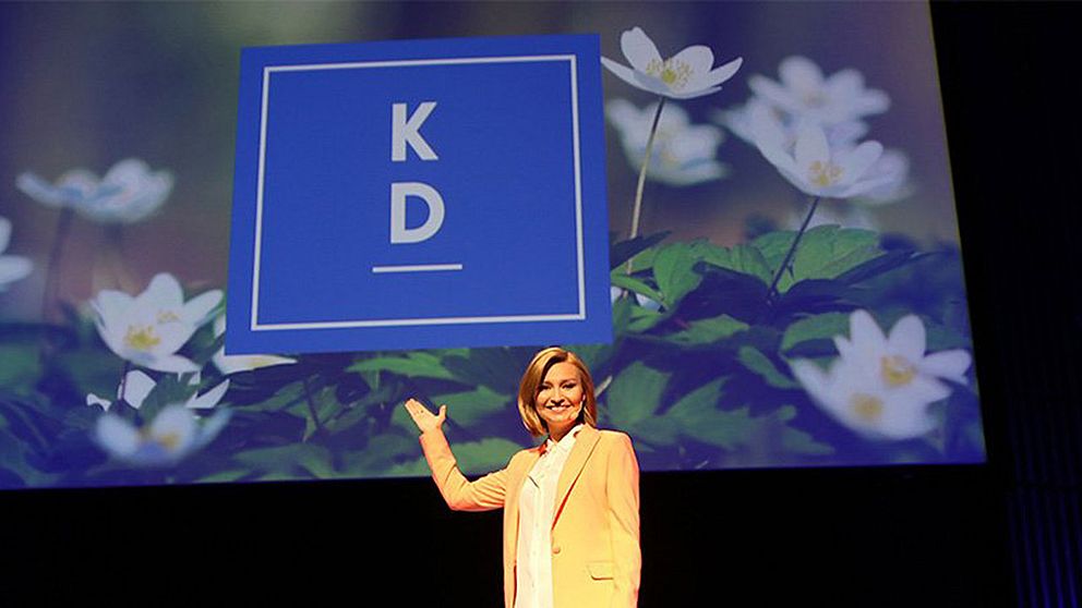 Ebba Busch Thor framför en bild på logotypen; En blå fyrkant med bokstäverna ”KD” ovanför ett streck.