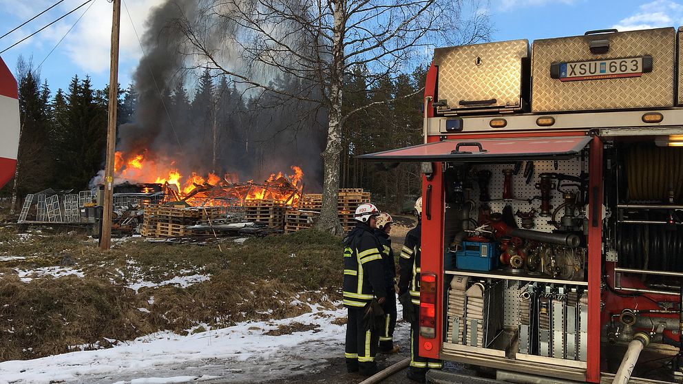 I förgrunden står personal från räddningstjänsten och i bakgrunden ser man ladugården som håller på att brinna ned.