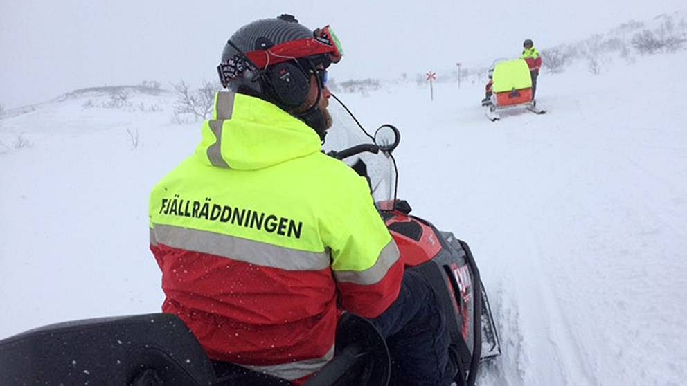 två män i fjällräddningens kläder med skoter och pulka på fjället