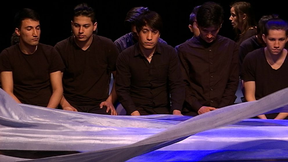 Fem unga killar sitter bredvid varandra på golvet med ett blått tygstycke som fladdrar i förgrunden. Scenen föreställer båtresan till Sverige över Medelhavet.