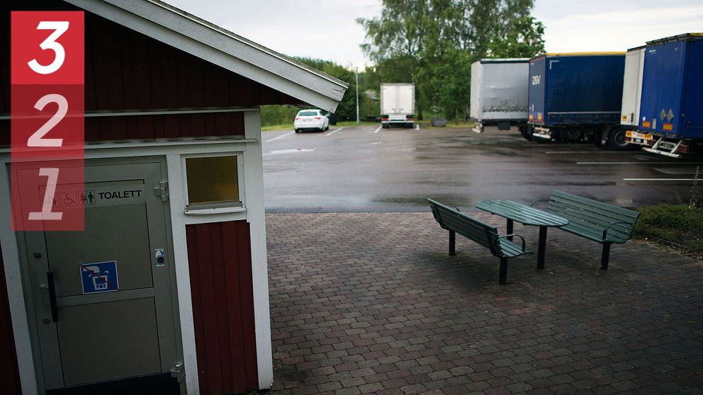 En parkeringsplats vid en rastplats med flera lastbilar parkerade. Till vänster i bilden finns en röd byggnad med en dörr märkt ”Toalett” för handikappanpassning. I förgrunden syns ett par bänkar och ett bord på en stenbelagd yta.