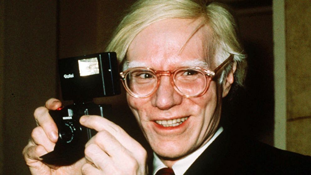 En leende Andy Warhol 1976, iklädd kostym och glasögon med en kamera i händerna.