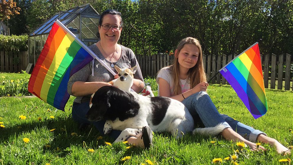 En kvinna och hennes dotter håller i regnbågsflaggor, de sitter i gräset med två hundar.