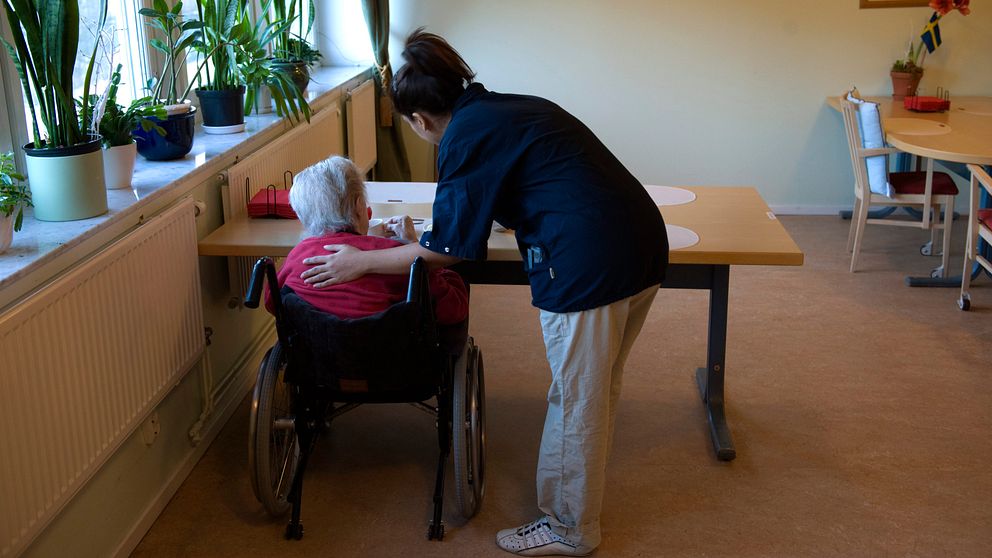Arkivbild med äldre person i rullstol och sköterska ståendes bredvid, båda med ryggen mot kameran