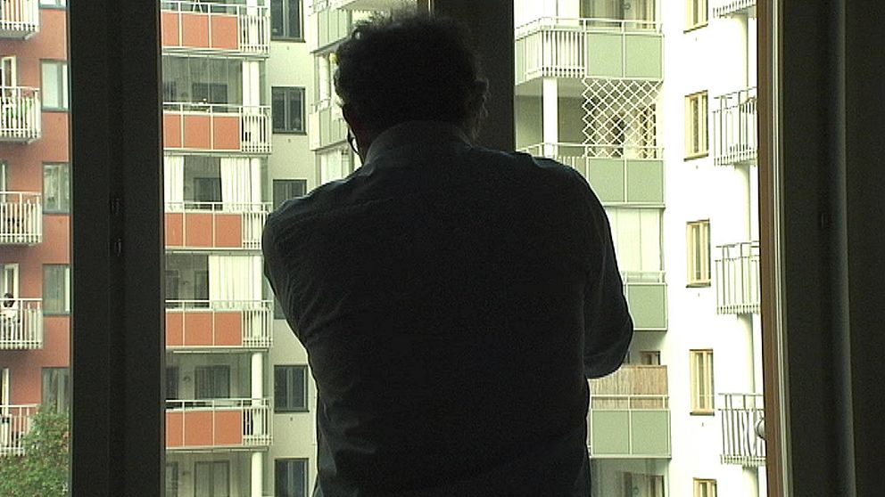 Anonym man står och tittar ut genom ett fönster i ett bostadsområde.