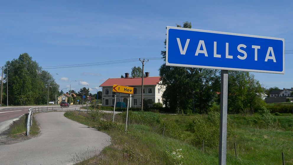 Vägskylt med ortsnamnet Vallsta till höger och en skymt av riksväg 83 till höger på bilden.