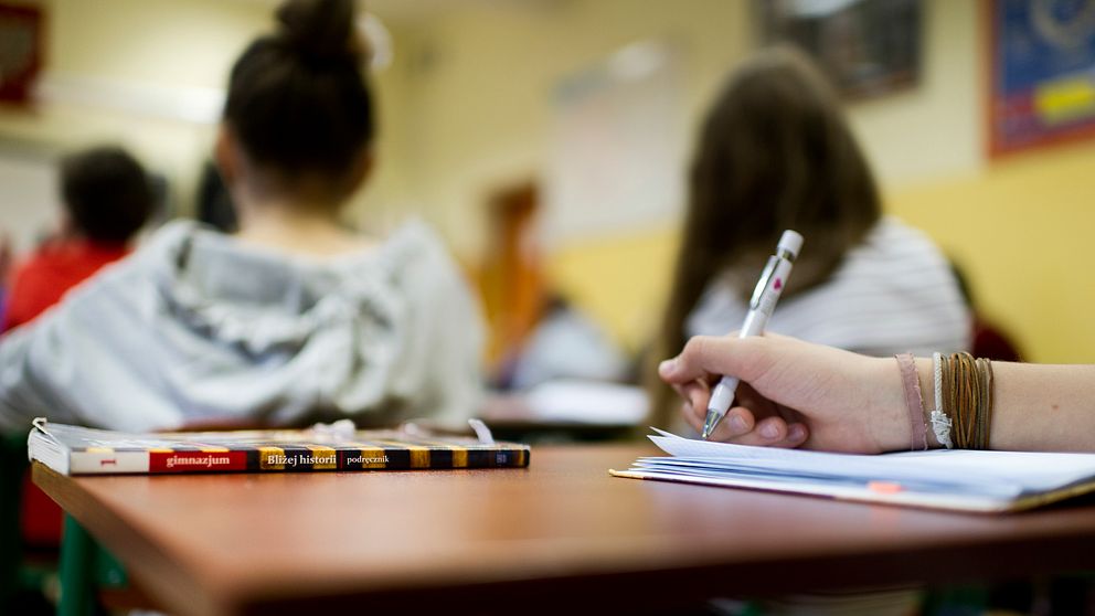 Elever ses bakifrån i klassrum, hand som skriver i förgrund