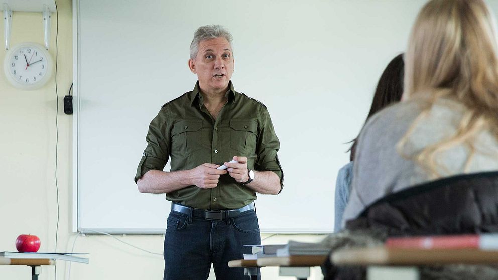 Man står och pratar i ett klassrum inför elever. En klocka syns i bakgrunden på väggen och på bänken framför honom ligger ett rött äpple.