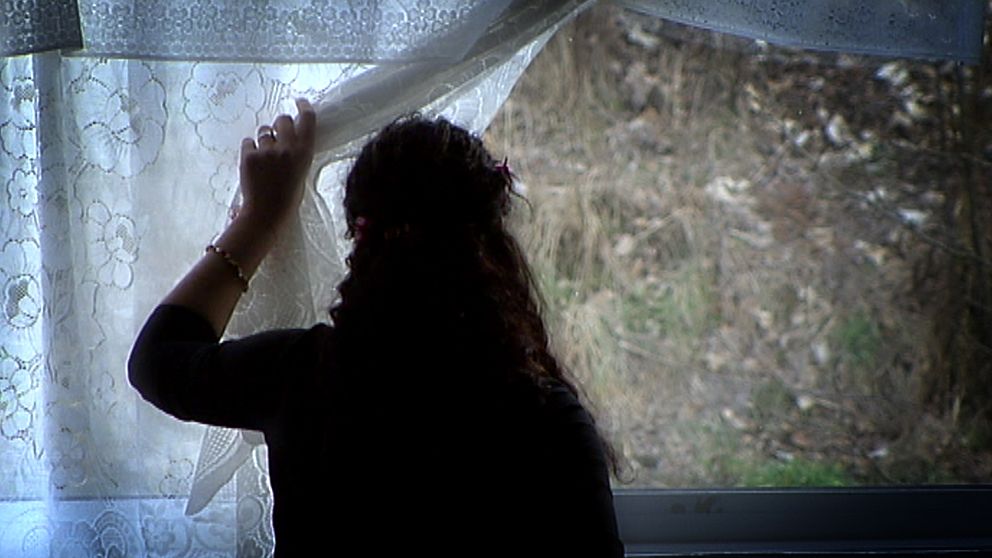 En person med långt hår står bortvänd mot kameran och tittar ut genom ett fönster med spetsgardiner.