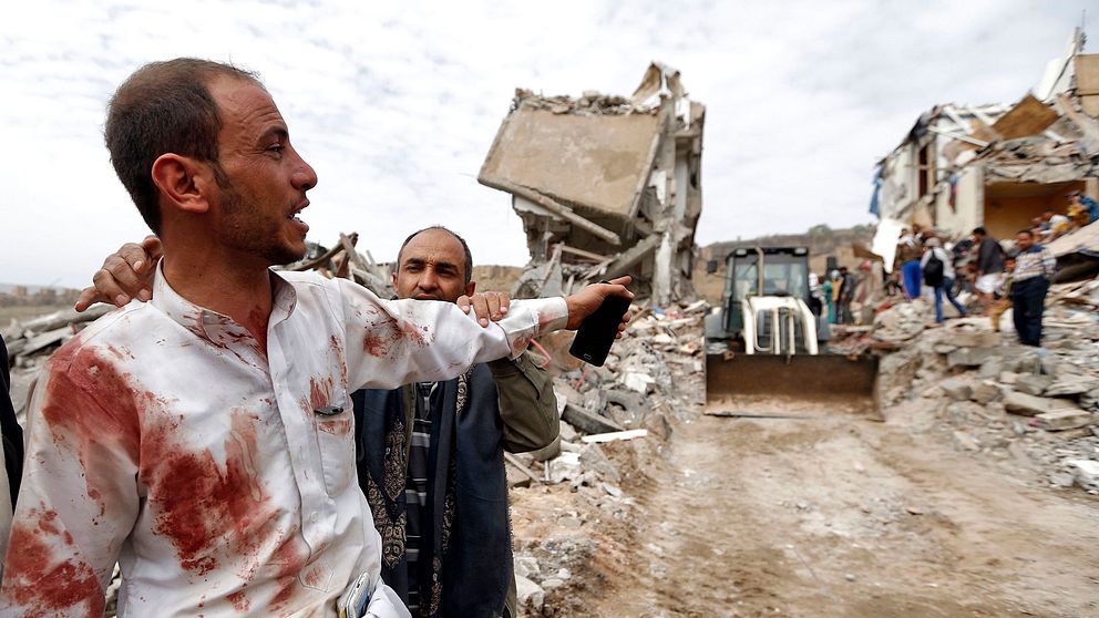 En man pekar mot en av de byggnader som förstördes i flygattacken, där man nu söker efter överlevande.