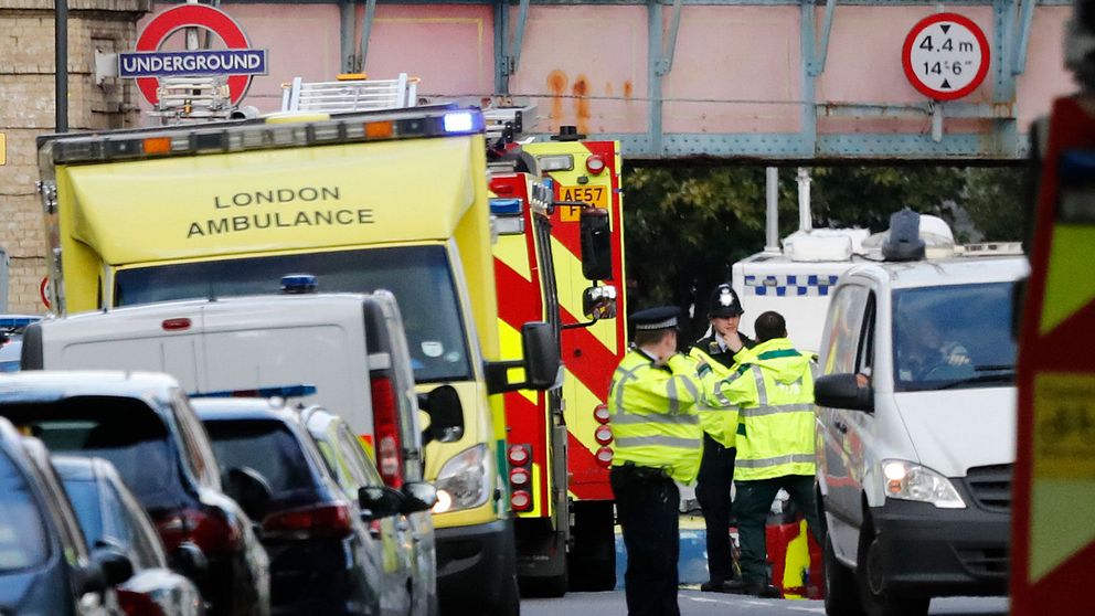 Tre polismän i gula jackor står och pratar med varandra intill en brandbil, en ambulans och en polisbil intill en bro vid en tunnelbaneskylt i London.