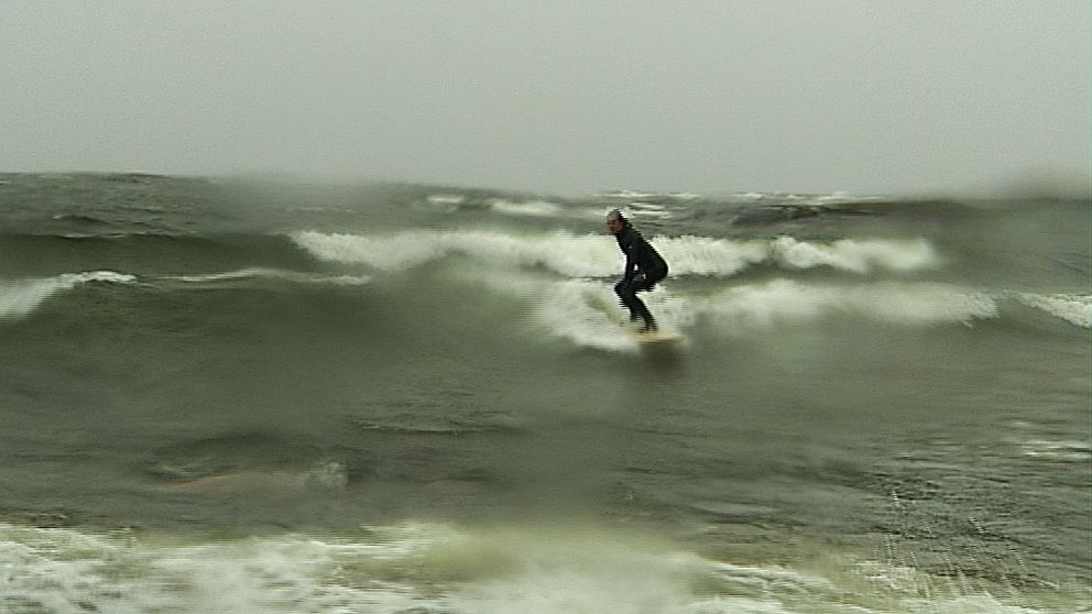 En man i svart dräkt står på en surfbröda ute i vattnet. Bilden är lite suddig av vattendroppar