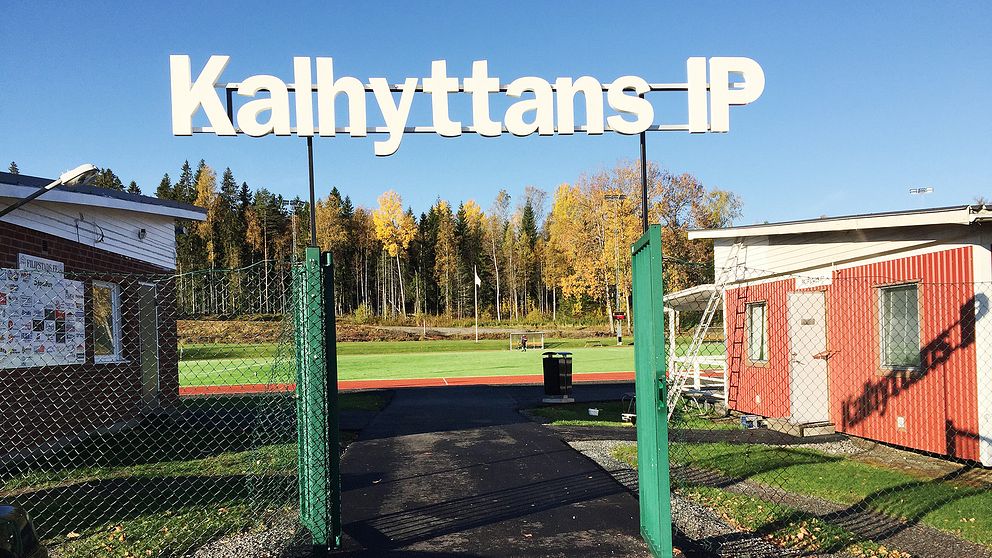 En grön grind står öppen, där syns träd och en gräsplan. En stor skylt i mitten av bilden ”Kalhyttans IP”.