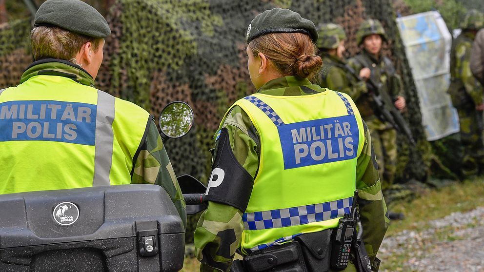 Två kvinnliga militärpoliser framför ett förband med soldater och ett kamoflerat militärfordon.