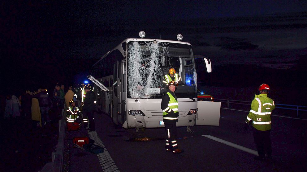 Vit buss med stora skador på framrutan. Räddningspersonal. Människor håller om varandra vid sidan av motorvägen.