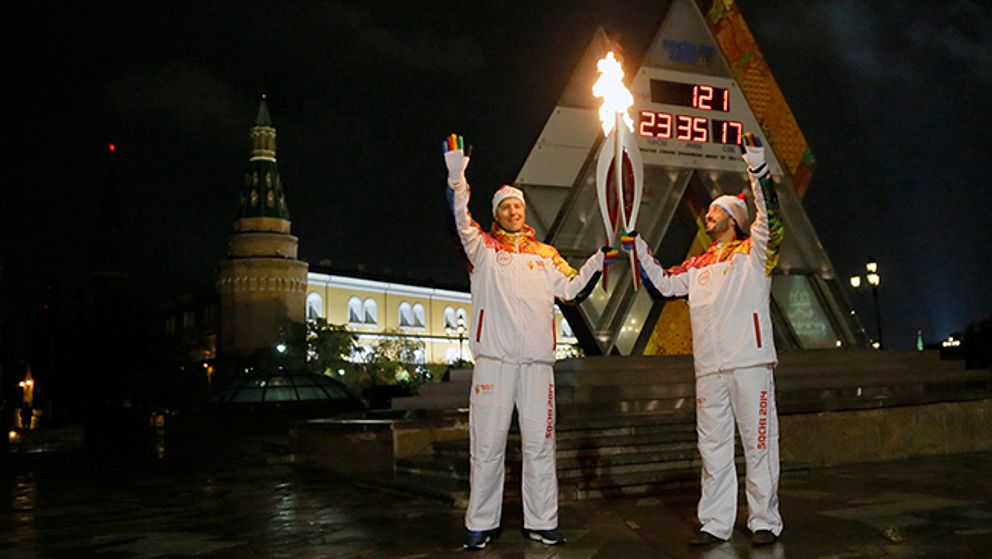 Den ryska handelsministern Denis Manturov och konståkaren Ilya Averbukh håller i olympiska facklor framför nedräkningen på Manegetorget i Moskva.