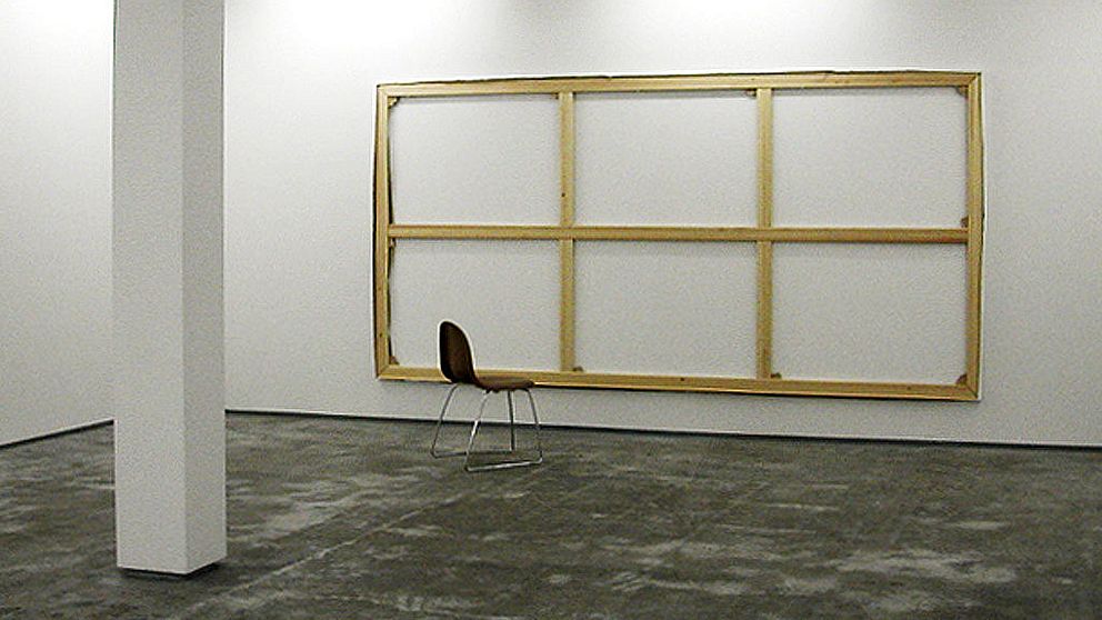 Ett vitt rum, på väggen hänger en stor träram, framför står en brun stol.