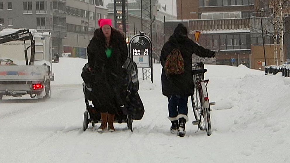 Två personer i svarta jackor går längs en väg där snön yr omkring dem.
