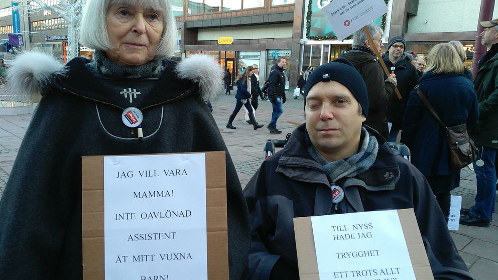 Birgitta och Erik står med varsin skylt på demonstrationen. ”Jag vill vara mamma, inte oavlönad assistent åt mitt vuxna barn.” ”Till nyss hade jag trygghet, ett trots allt hyfsat liv. Och nu?”