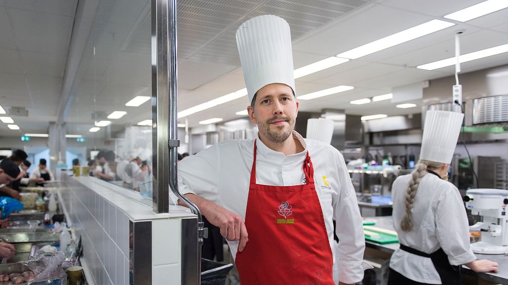 Daniel Roos står i ett kök med massa kockar som arbetar bakom. Han har på sig vit rockmössa, vit kockskjorta och rött förkläde.