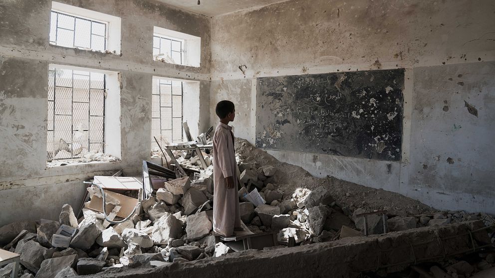 En ensam pojke står i en hög av ruiner av det som en gång var ett klassrum.